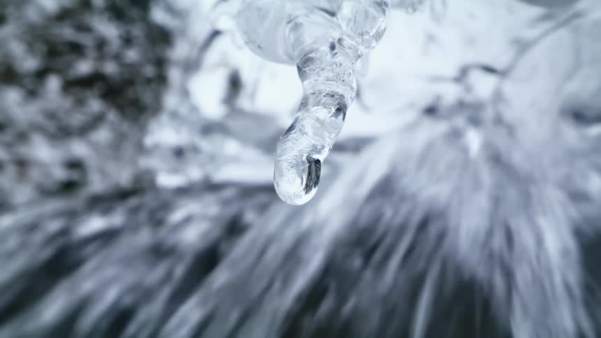 Water drop dripping from an icicle hanging near a waterfall at winter