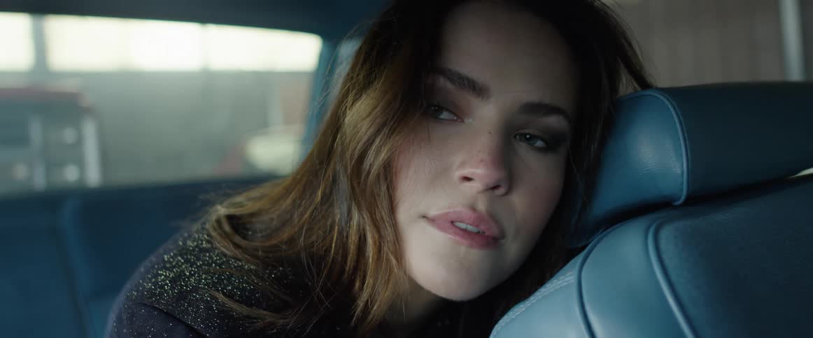 A woman sitting in the back seat of a car leans against the front seat provocatively