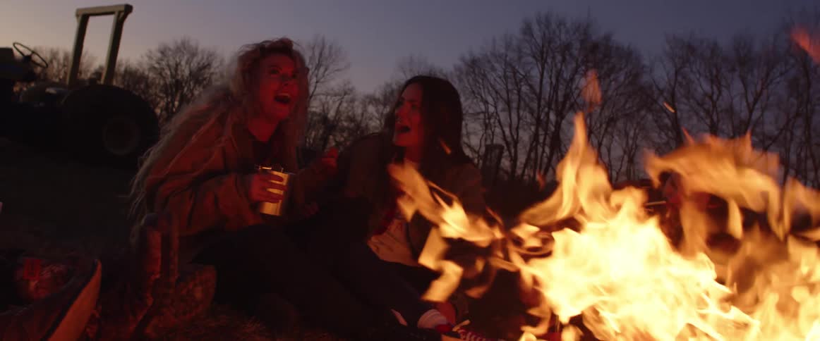 Group of friends talking and laughing sitting near to the raging fire on a campfire at sunset