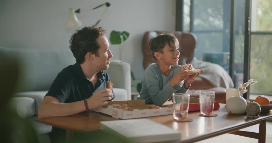 Father and son talk while sitting in living room eating pizza