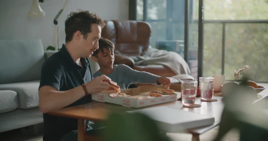 A man and a boy sitting on the floor next to a table with a box of pizza