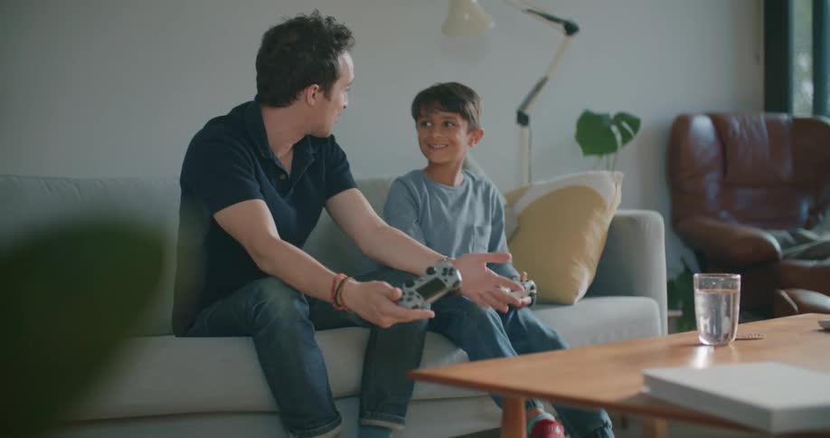 A man and boy play video games while sitting together on a sofa in the living room of a house during the day
