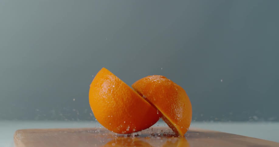 An orange separates in half and water sprays from it as it is dropped on to a cutting board