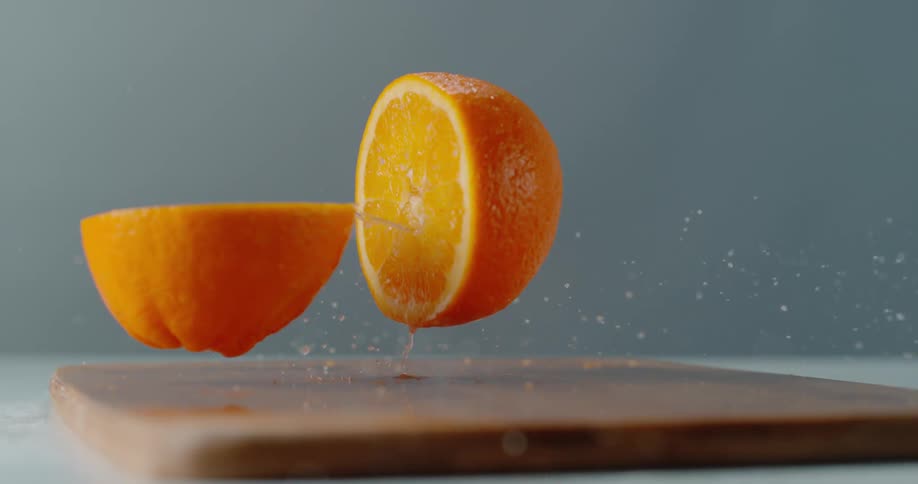 An orange falls down on a cutting board and splits in half where it had been cut and juice squirts out