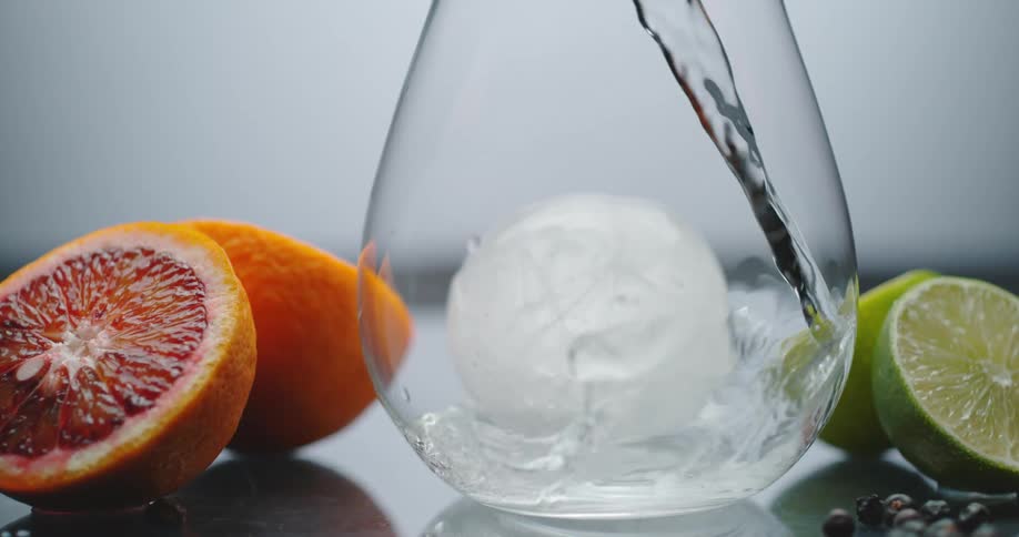Water pours into a clear glass with an ice ball inside and orange and lime on either side