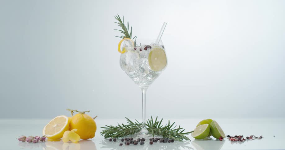 An ice cube falls into a full drinking glass adorned with fruit causing the contents to splash into the air