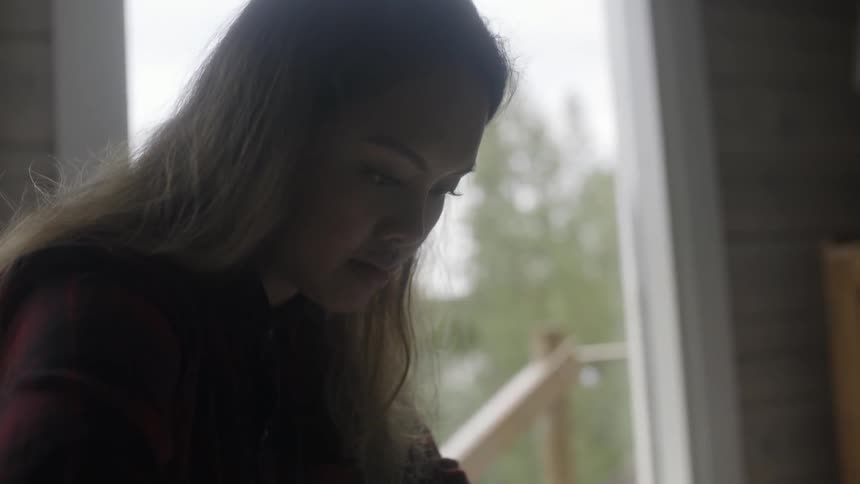 A woman with long hair looks down in front of a window