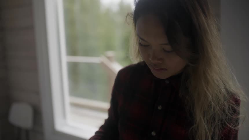 An asian woman standing in a house during the day works on something below camera view