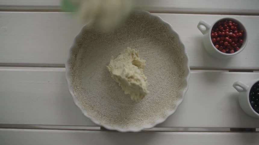 Pie crust sits on table next to a cup of berries, and a person places a dollop of filling in the crust