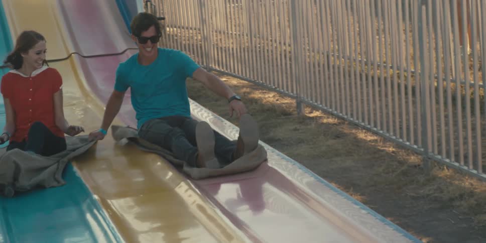A young woman and man slide side by side down a large slide in an amusement park atop blankets