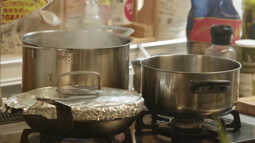Steam emerges from a sauce pan on a stove top holding several pans