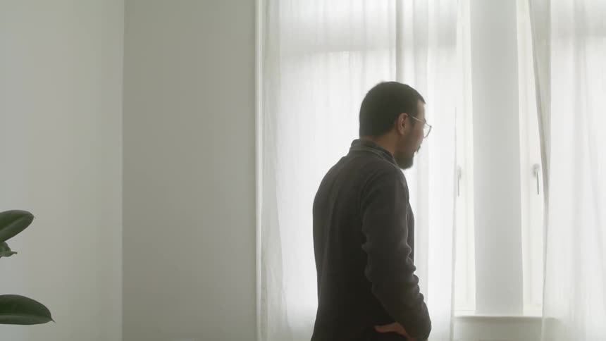 A man laughs while speaking on his smartphone as he pulls aside a curtain to glance outside the window