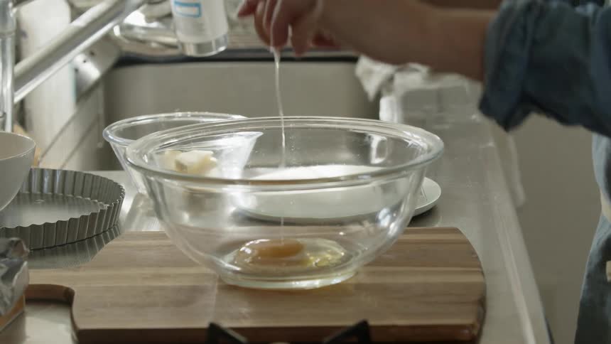 Woman with blue shirt cracks egg into glass bowl in the kitchen