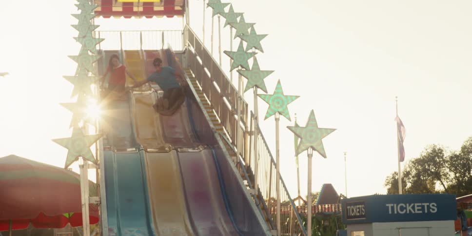 A man and woman hold onto burlap sacks and each other as they come down a tall slide at a carnival