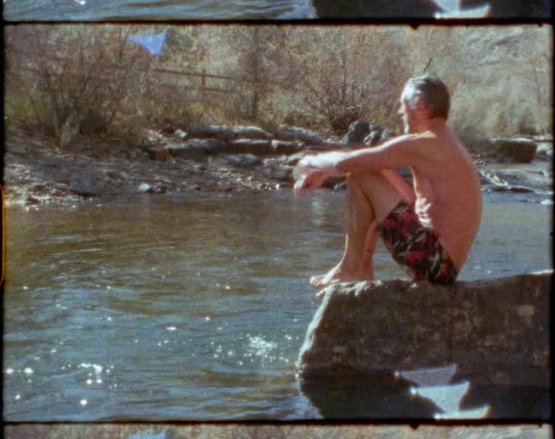 Man with swimming shorts sits on rock next to the river