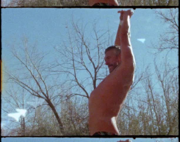 A barechested man in swim trunks stands near a line of trees and waves his arms around