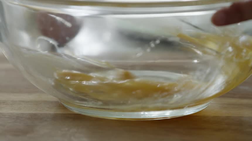 A hand holds a clear glass bowl steady as a person whisks an egg
