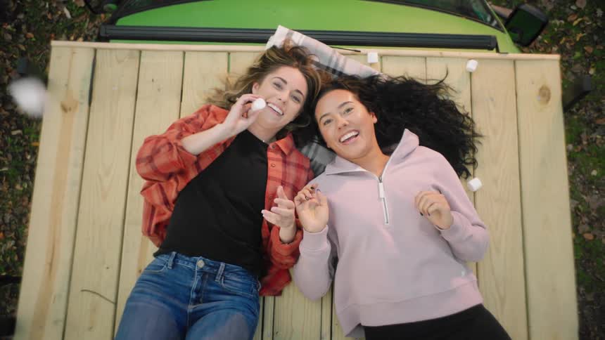 Two female friends laying on a wooden pallet over the roof of a car playing with marshmallows
