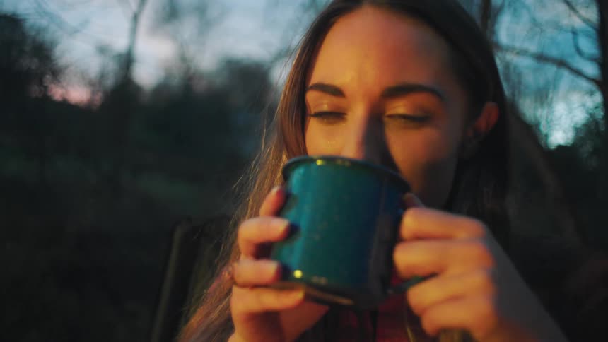 Woman taking a drink from enamelware mug while camping at sunset
