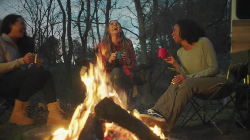 Group of female friends talking and laughing while drinking hot beverages in front of a campfire