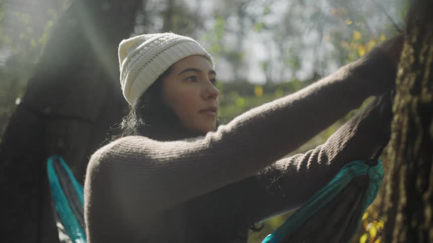 A woman with a hat hanging up a hammock on a tree with the sky and clouds in the background