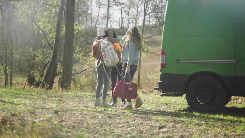 Three women hugging each other next to a vehicle with trees the sky and clouds in the background