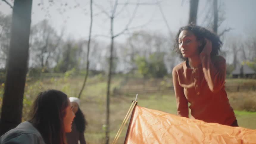 Three female friends are putting up an orange tent at a campsite