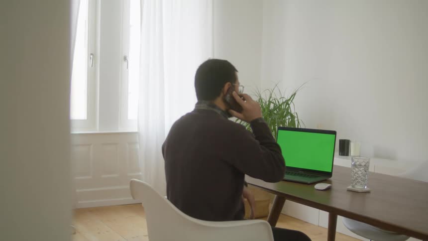 A man with a smart phone sitting on a chair at a table with a laptop