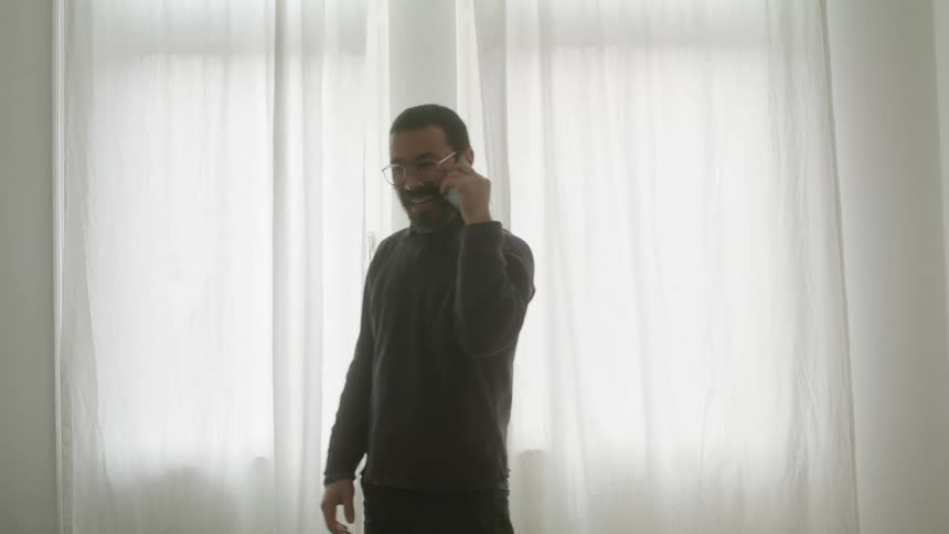 A man with glasses in a room talking on a smart phone with windows and curtains in the background