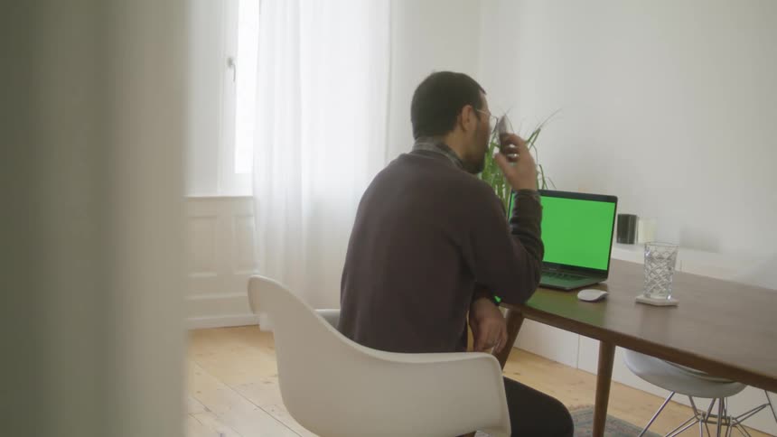 Man sitting in a desk at home answer a phone call on his smartphone while working on a laptop