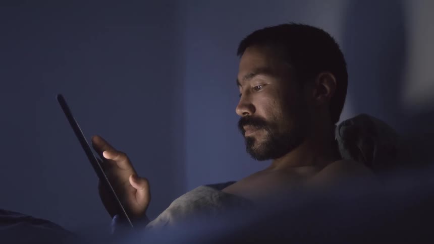 A man uses a finger to swipe the screen of a laptop as he lies in bed in a dark room