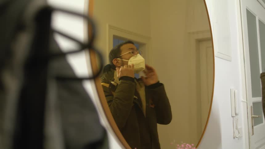 A man puts on a padded winter coat and looks in a mirror to put on a face mask before leaving his house
