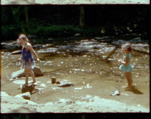 Two girls next to a river with water and rocks with trees and plants in the background