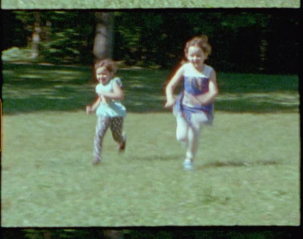 A pair of young girls run together across a field of grass on a sunny day