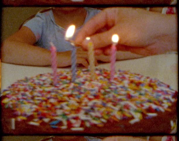 An adult lights four candles on a chocolate birthday cake as a pair of girls sit and wait