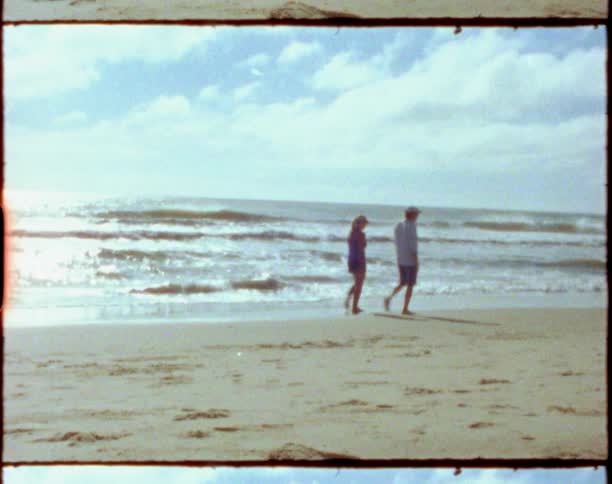 A man and woman walk together along a beach just out of reach of the waves on a sunny day