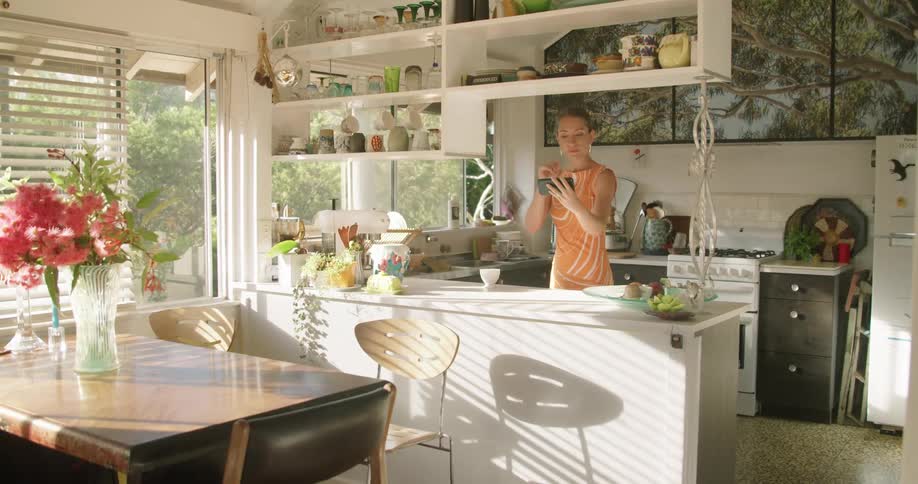 Woman wearing peach colored dress sets down coffee cup to wave at phone she is video calling on