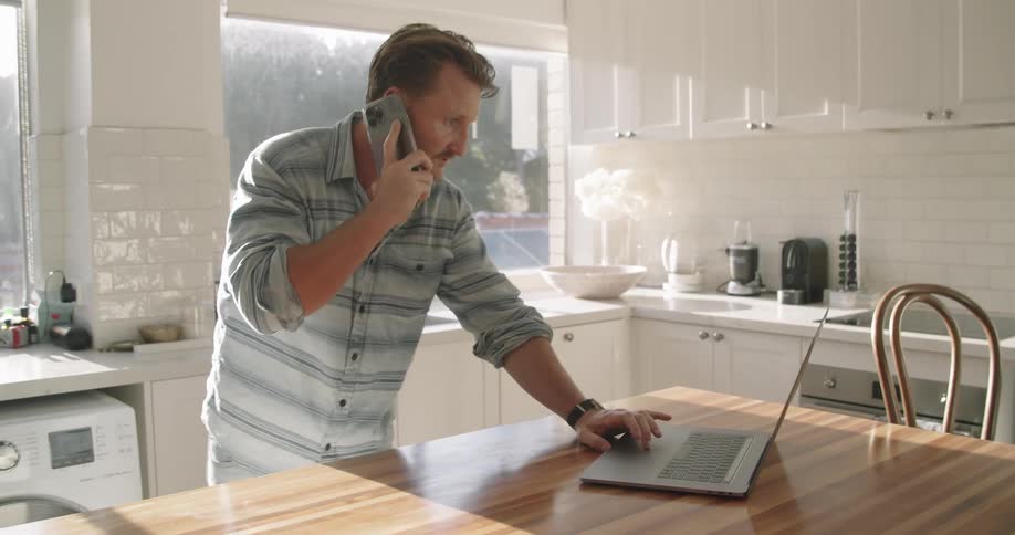 Man standing in kitchen holds phone to his ear while bending to look at laptop screen