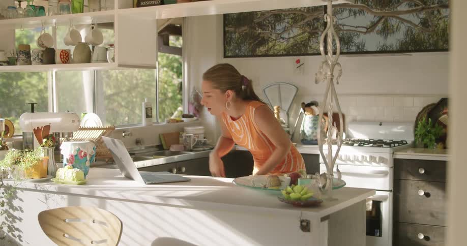 A woman makes a hand gesture towards her laptop computer in a video chat on top of a kitchen counter at daytime