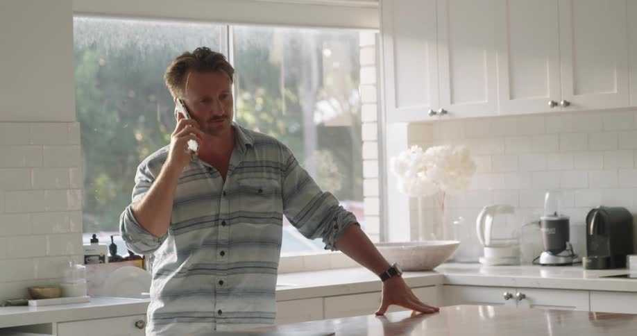 A man rests his arm on the counter of his all white kitchen while talking on the phone