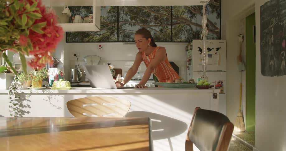 A woman leans back and forward while standing in her kitchen and using a laptop