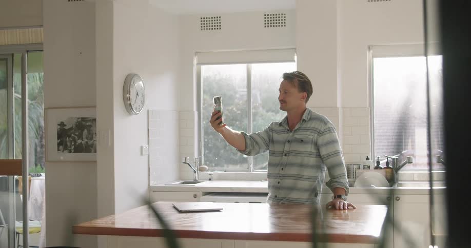 A man stands in his kitchen and waves at his cell phone as he video chats
