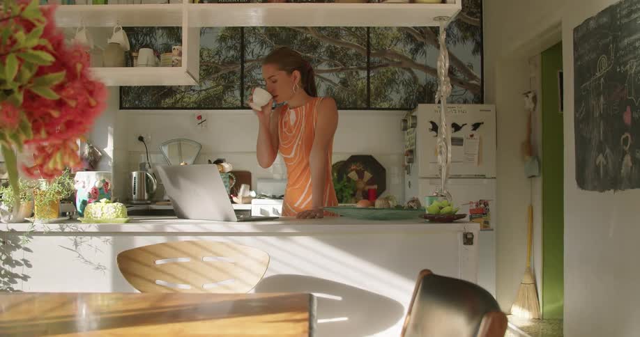 A woman stands in her kitchen and drinks coffee as she uses a laptop