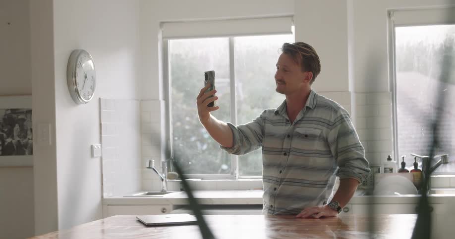 A man waves to someone during a video call on his smartphone as he stands and talks in the kitchen