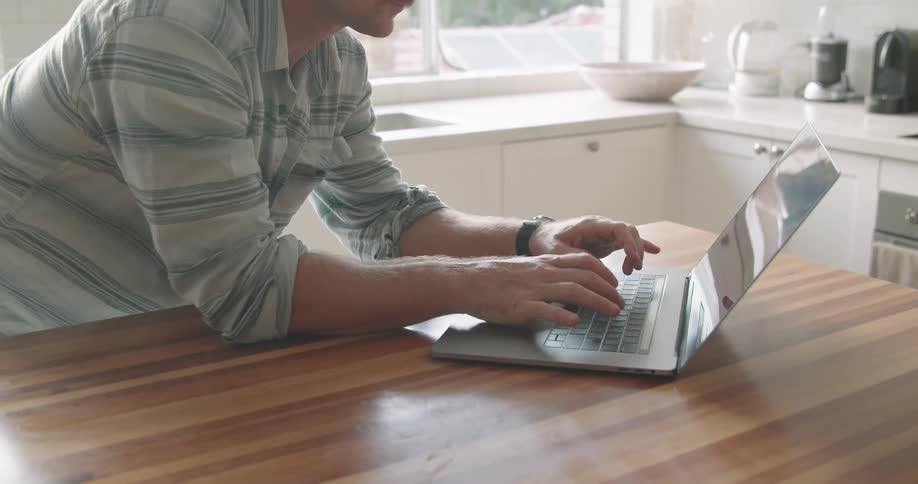 A man typing on a laptop while leaning over a kitchen counter