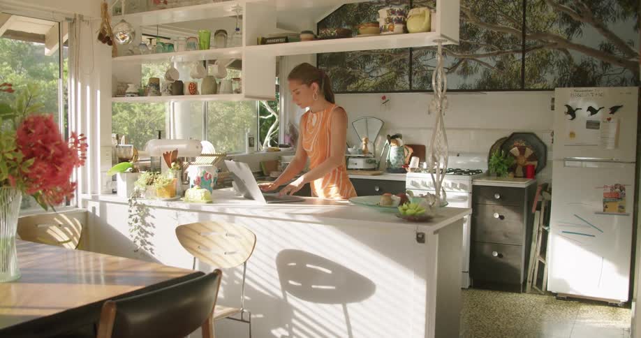 A woman stands in her kitchen and drinks coffee and looks at her laptop