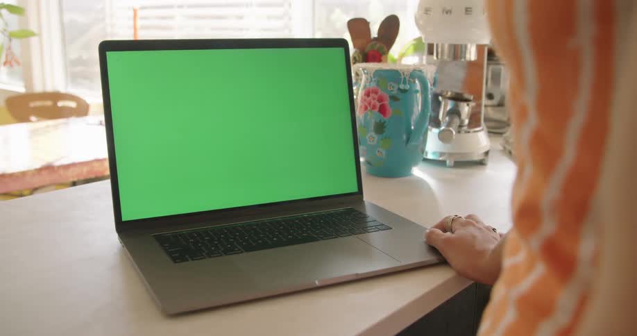 A woman stands at her kitchen counter and looks at a green screen on her laptop computer