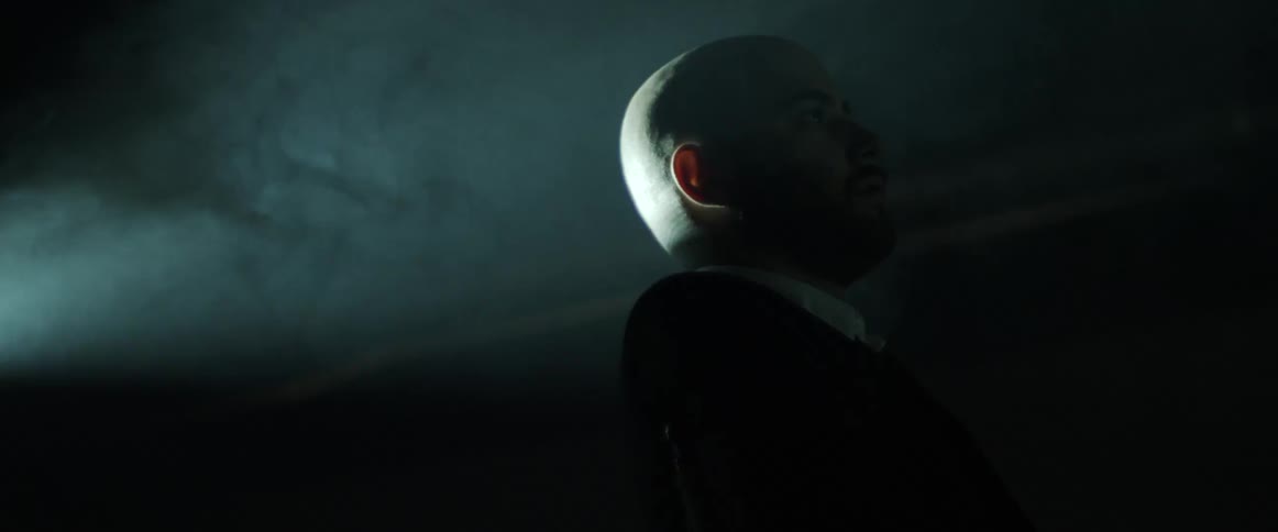 A bright light shines on the back of a bald man standing outside at night as smoke billows around him