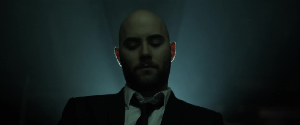 A man in a black suit and tie looks up as he stands in front of a bright light