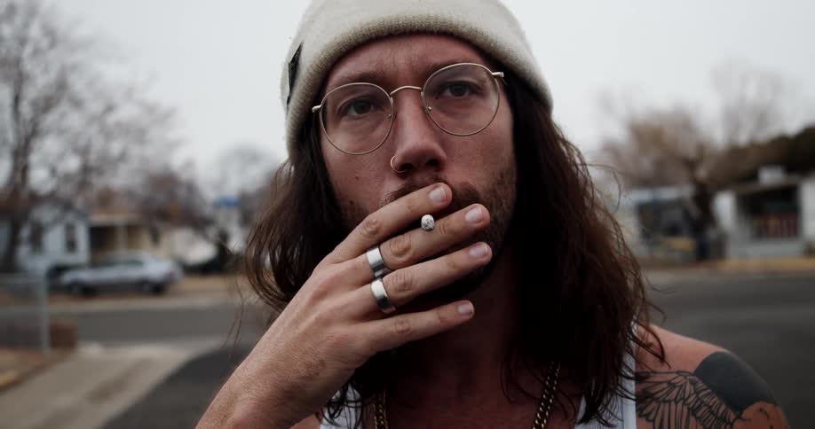 A man tattooed man smokes a cigarette while standing in the road outdoors in a residential neighborhood during the day